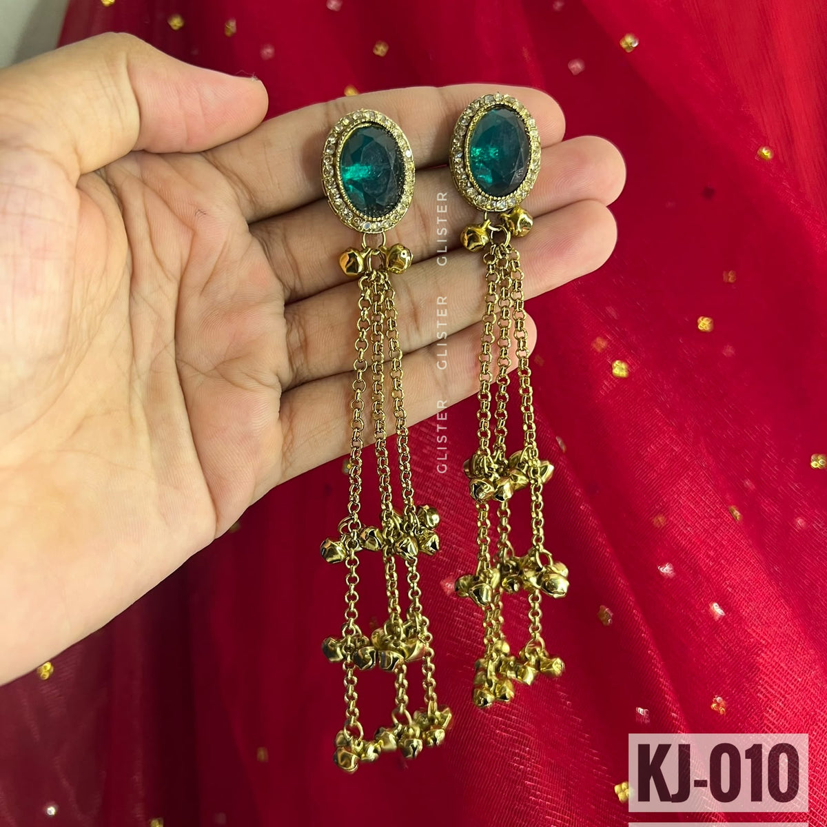 Glister KJ-010: gold-tone long earrings with oval deep green accents, clear decorative borders, hanging chains and bell-like drops, held against red fabric.