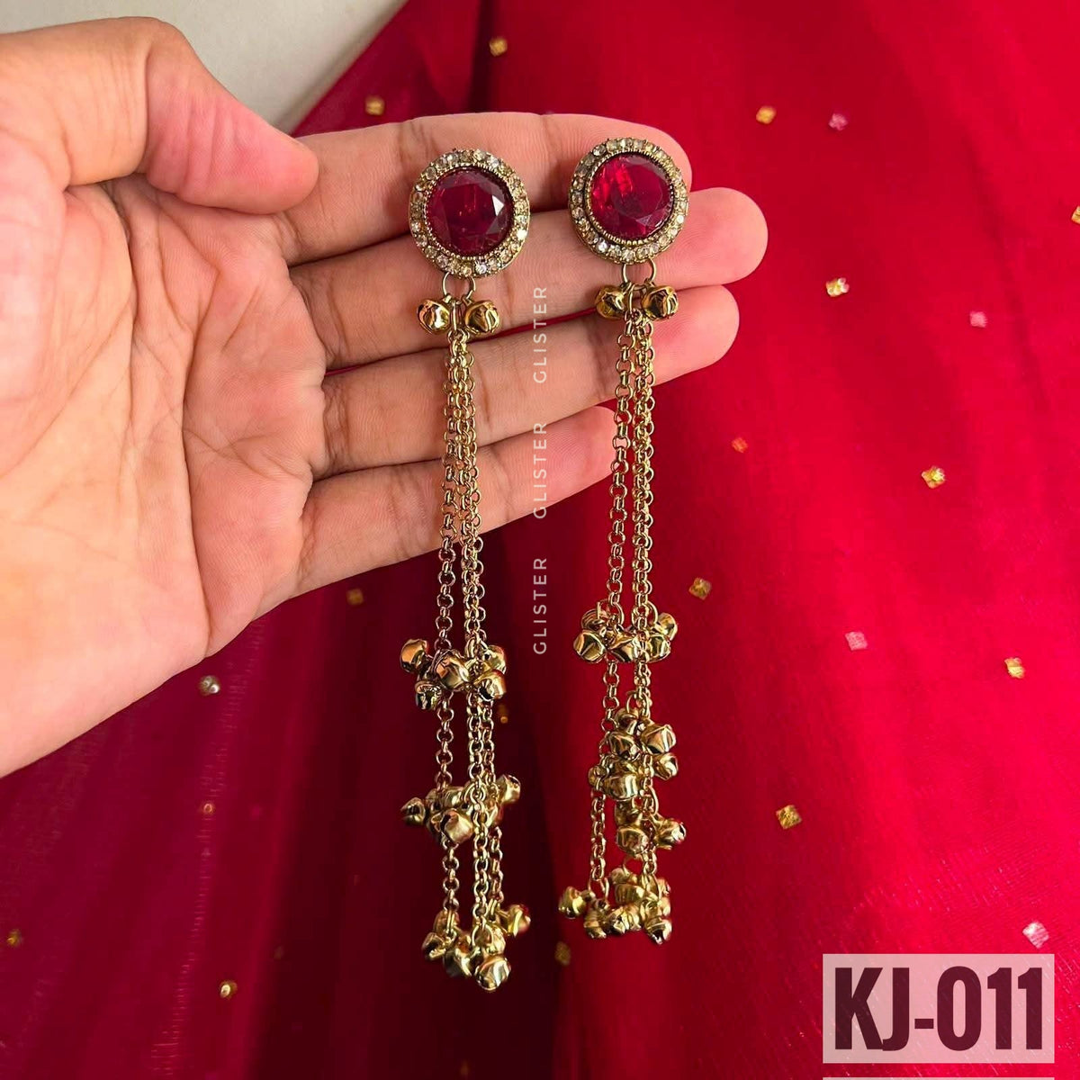 Glister KJ-011: gold-tone long earrings with oval deep pink accents, clear decorative borders, hanging chains and bell-like drops, held against red fabric.