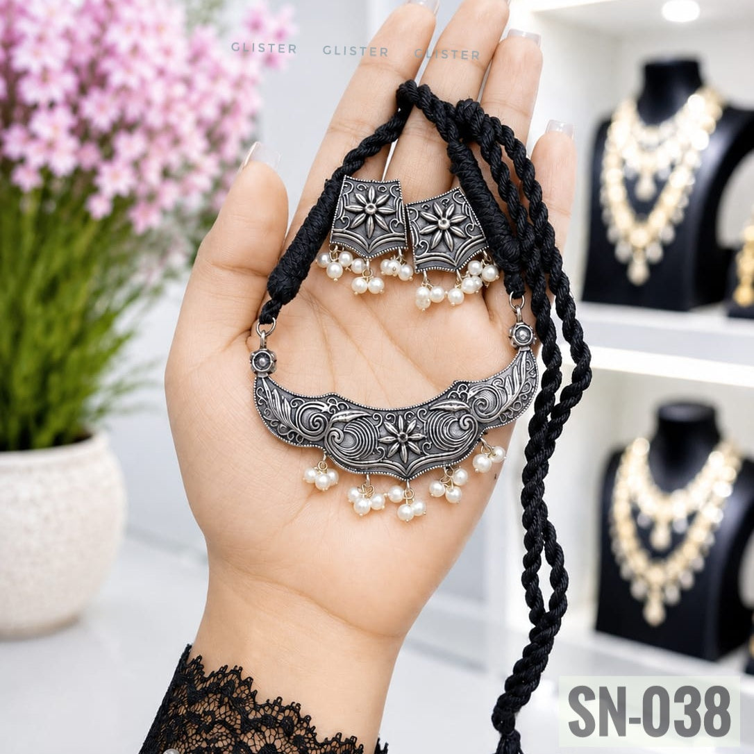 Glister SN-038: A curved floral pendant on a black braided cord, paired with square floral earrings and white bead-like drops.
