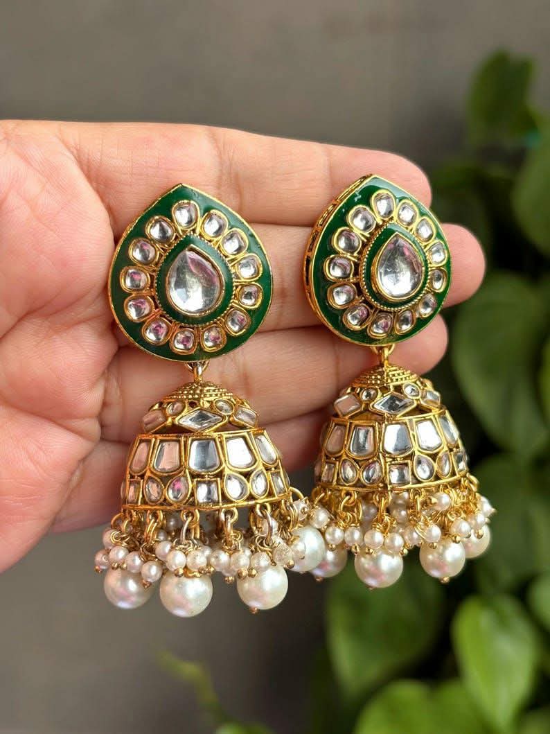 Green and gold-tone jhumka earrings with white bead drops, held in a hand